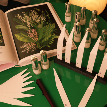 Sample perfume bottles with blotters on a green surface next to an open book with botanical illustrations.